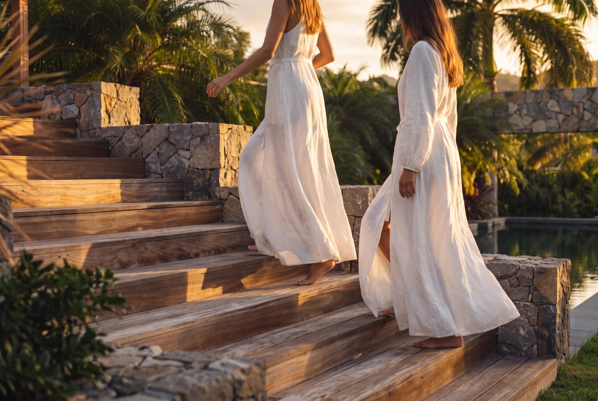 Two guests in white going up the wooden steps at sunset, the stone walls and the pool beside them