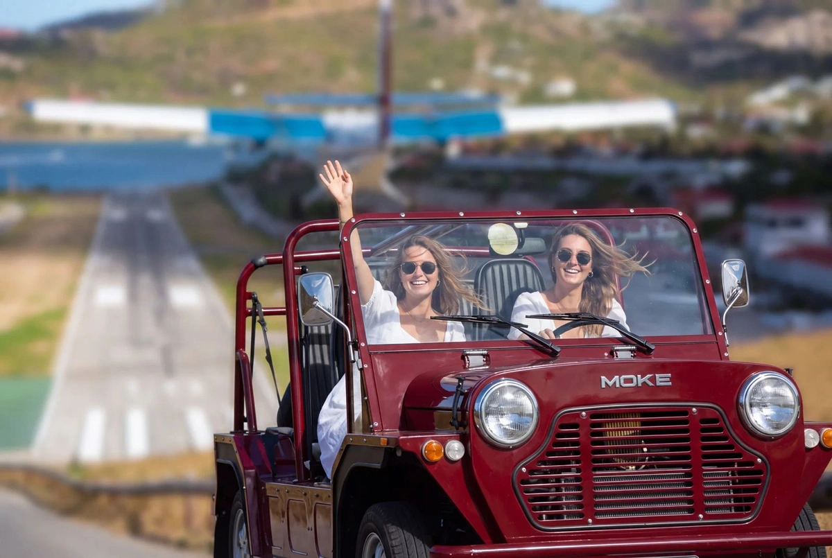 Two guests waving from a red Moke, the airstrip and the hills behind