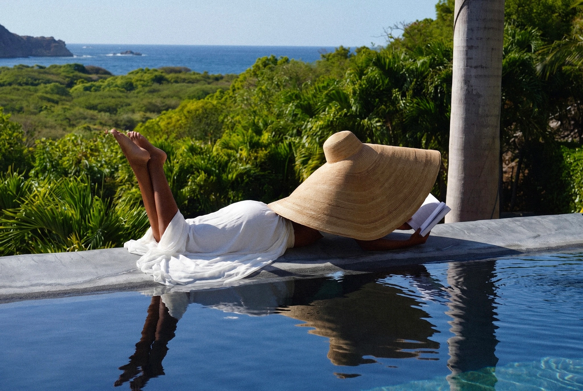 Reading at the pool's edge under a wide straw hat, the bay and the headland beyond